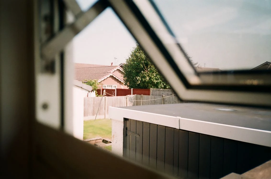 Fenêtre entrouverte sur un jardin, une clôture en bois et des maisons voisines